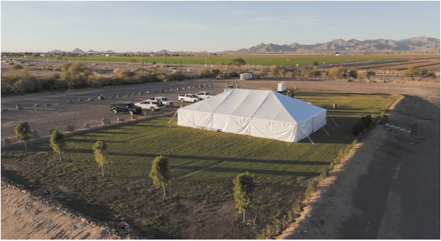 Aerial view of a large white tent with vehicles parked nearby, set in a grassy area with a backdrop of mountains.