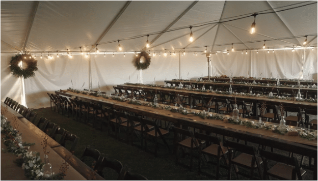 Elegant event setup under a tent with string lights, long tables, chairs, and floral decorations.