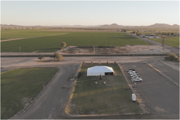 Aerial view of a rural setting with a large tent, vehicles, and vast farmlands in the background.