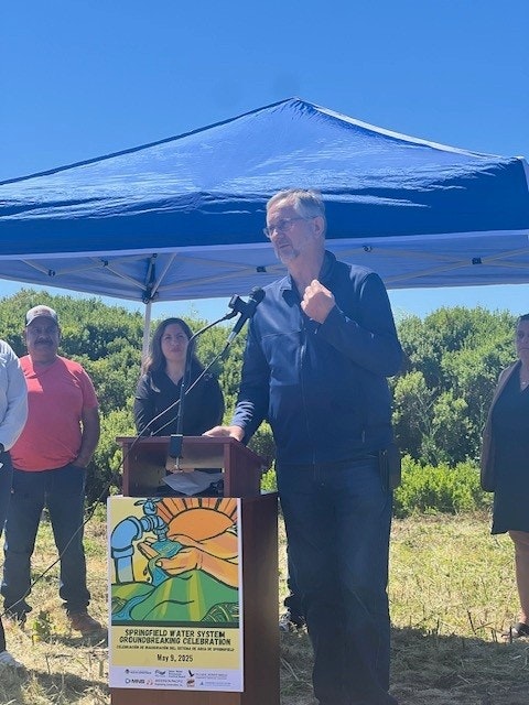 A person speaks at a podium under a blue canopy with others around, and a poster about Springfield Water System is displayed.