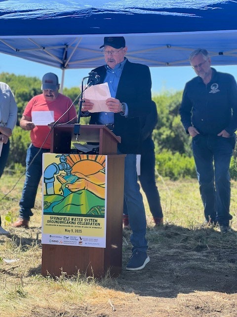 A man speaks at a podium under a blue canopy for a "Springfield Water System Groundbreaking Celebration" event. Others stand nearby.