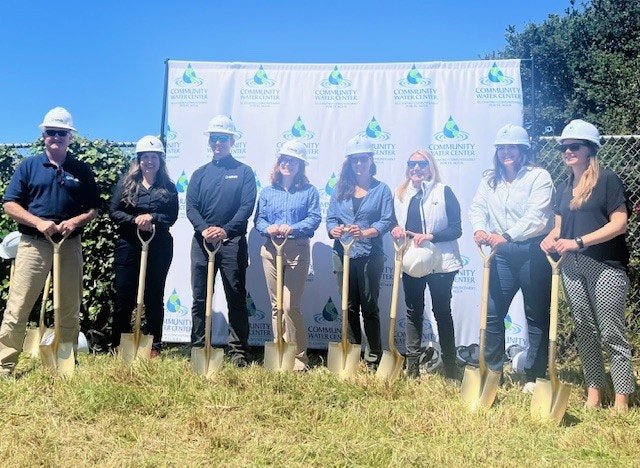 A group of people in hard hats holding shovels stand in front of a "Community Aquatic Center" backdrop on grass.