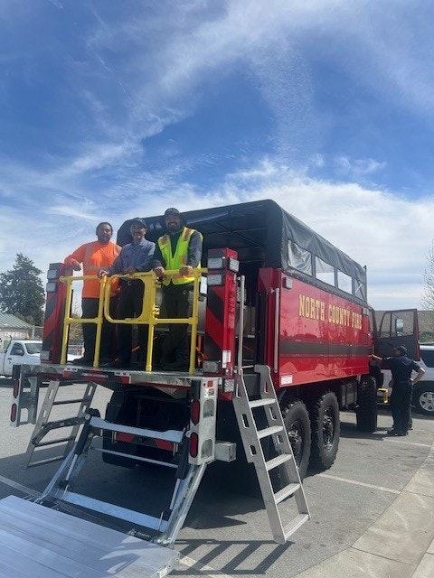 A red fire truck with "North County Fire" written on it, featuring workers on the rear platform in a parking lot.
