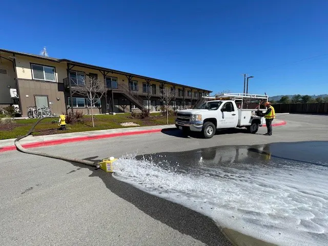 A white truck is spraying water and foam onto a parking lot near a multi-unit building on a clear day.