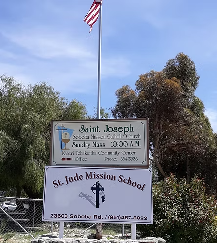 Signs for Saint Joseph's Church and St. Jude Mission School, with contact info and a flagpole, surrounded by trees.