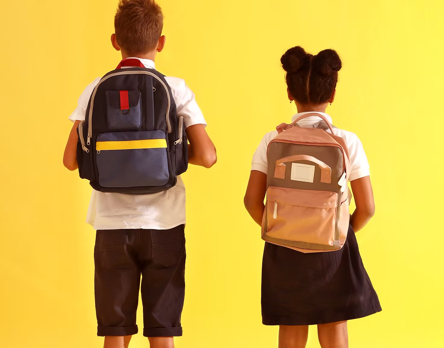 Two children with backpacks stand against a yellow background, viewed from behind.