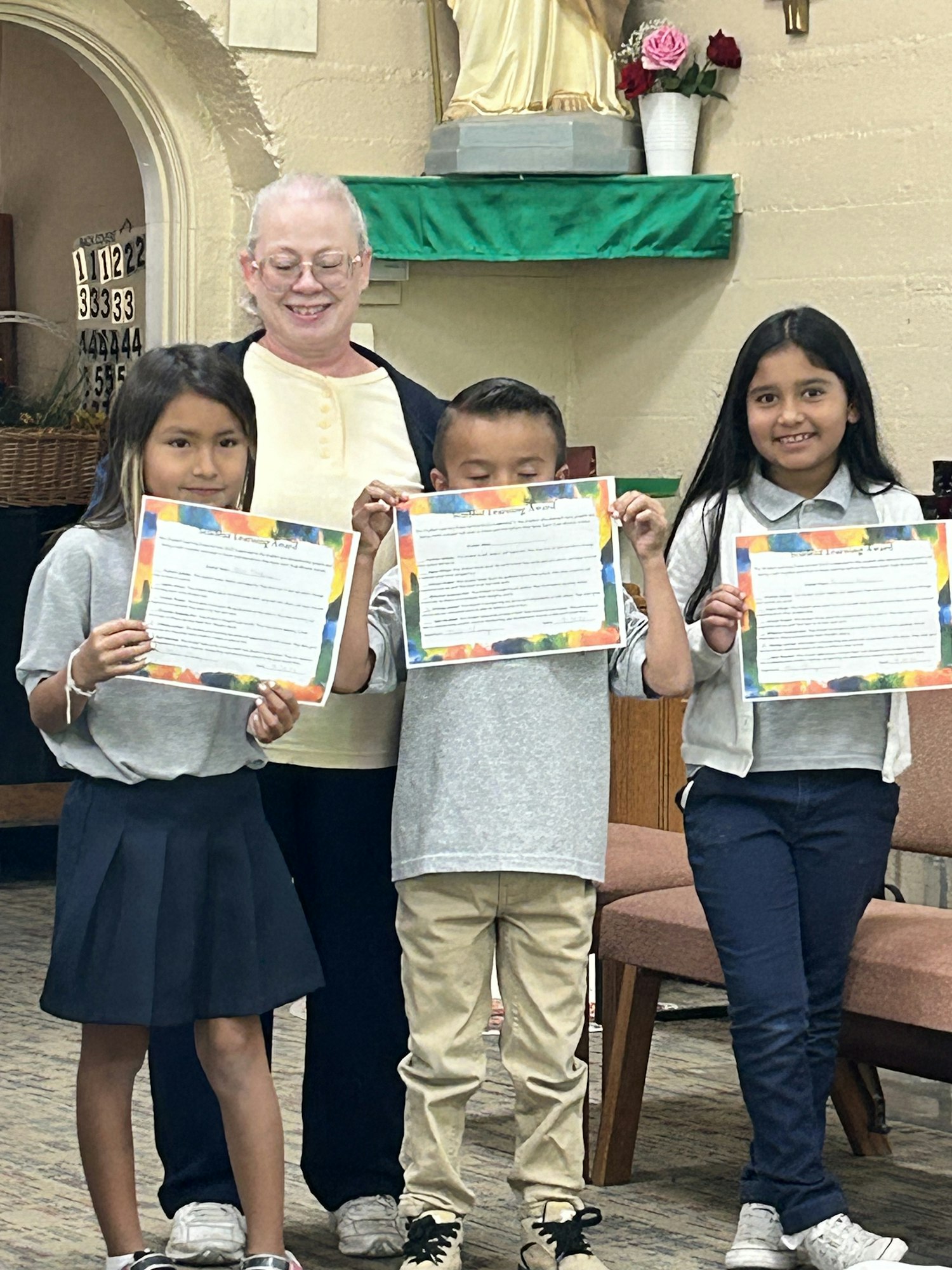 Four people holding colorful certificates, smiling inside a room with religious decor.