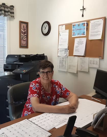 A person sitting at a desk in an office with calendars, a bulletin board, and a printer in the background.