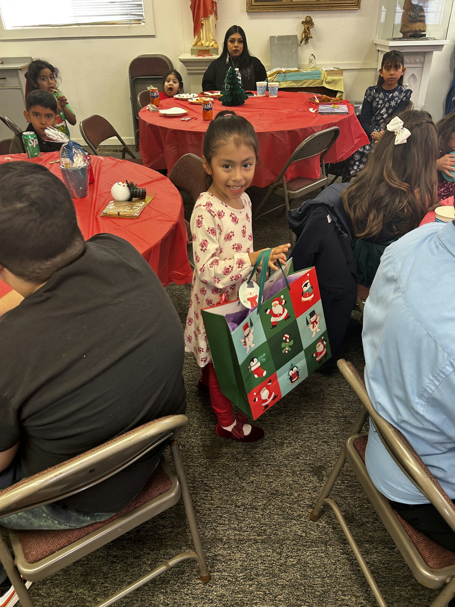 A festive gathering with children, red tablecloths, and a young girl holding a decorated gift bag, smiling joyfully.