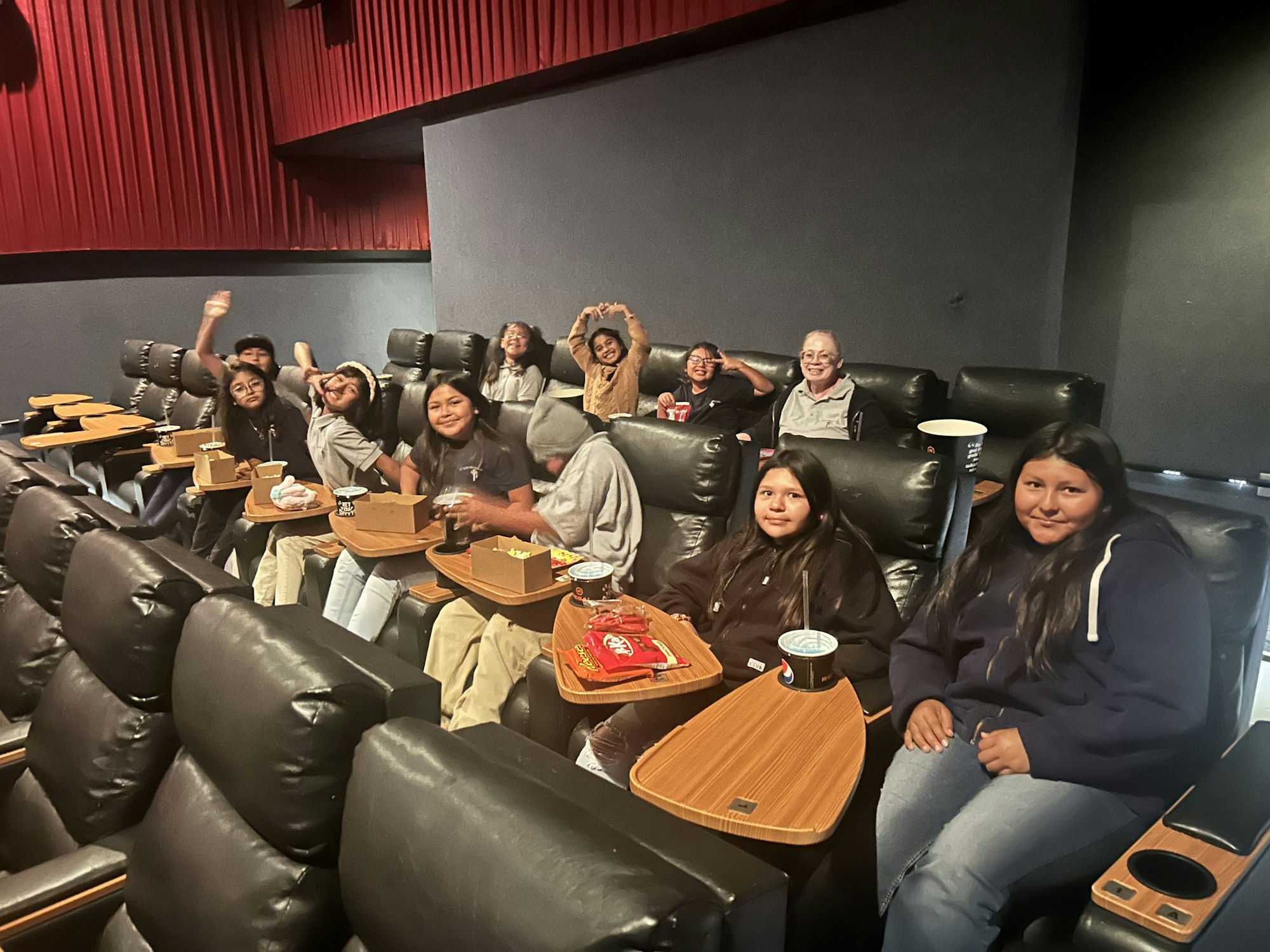 A group of people sitting in a theater, smiling and enjoying snacks, with leather recliner seats and wooden tables.