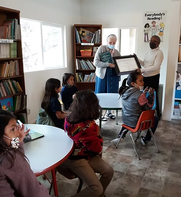 A group of children sit around tables in a library. Two adults are standing, one holding a certificate.