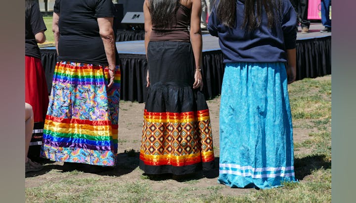 Three people in colorful skirts stand outdoors in front of a stage.