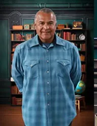 The image shows a man wearing a blue checkered shirt, standing in front of a bookshelf with various items.