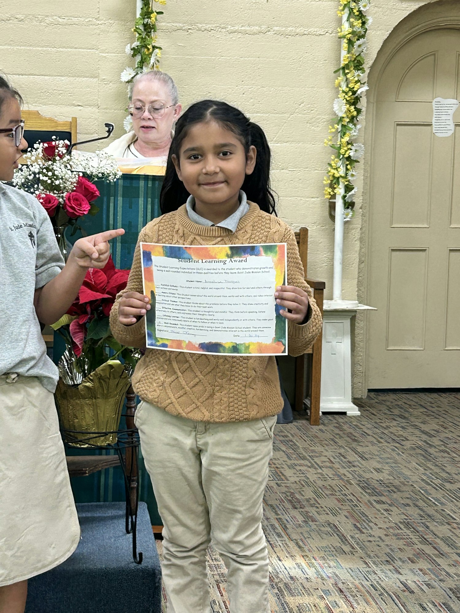 A child holding a "Student Learning Award" certificate, smiling in front of an adult, with flowers in the background.