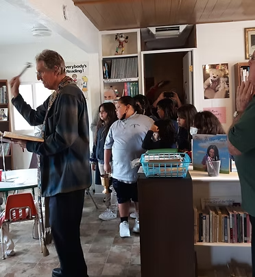 A person appears to conduct a ritual or ceremony in a room with several children and adults, surrounded by books and posters.