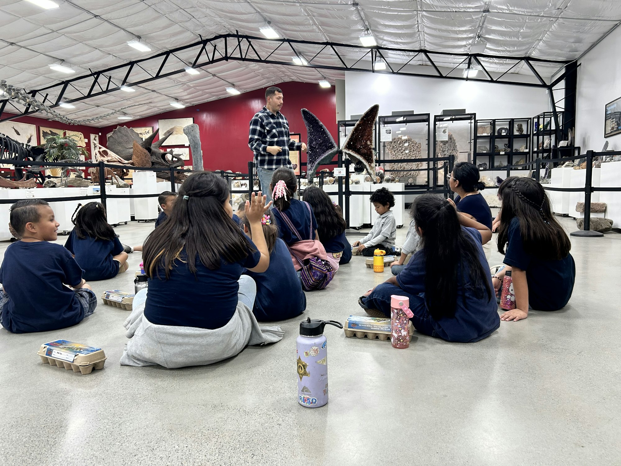 A group of children sit on a museum floor, listening to a guide. Dinosaur skeletons are displayed around.