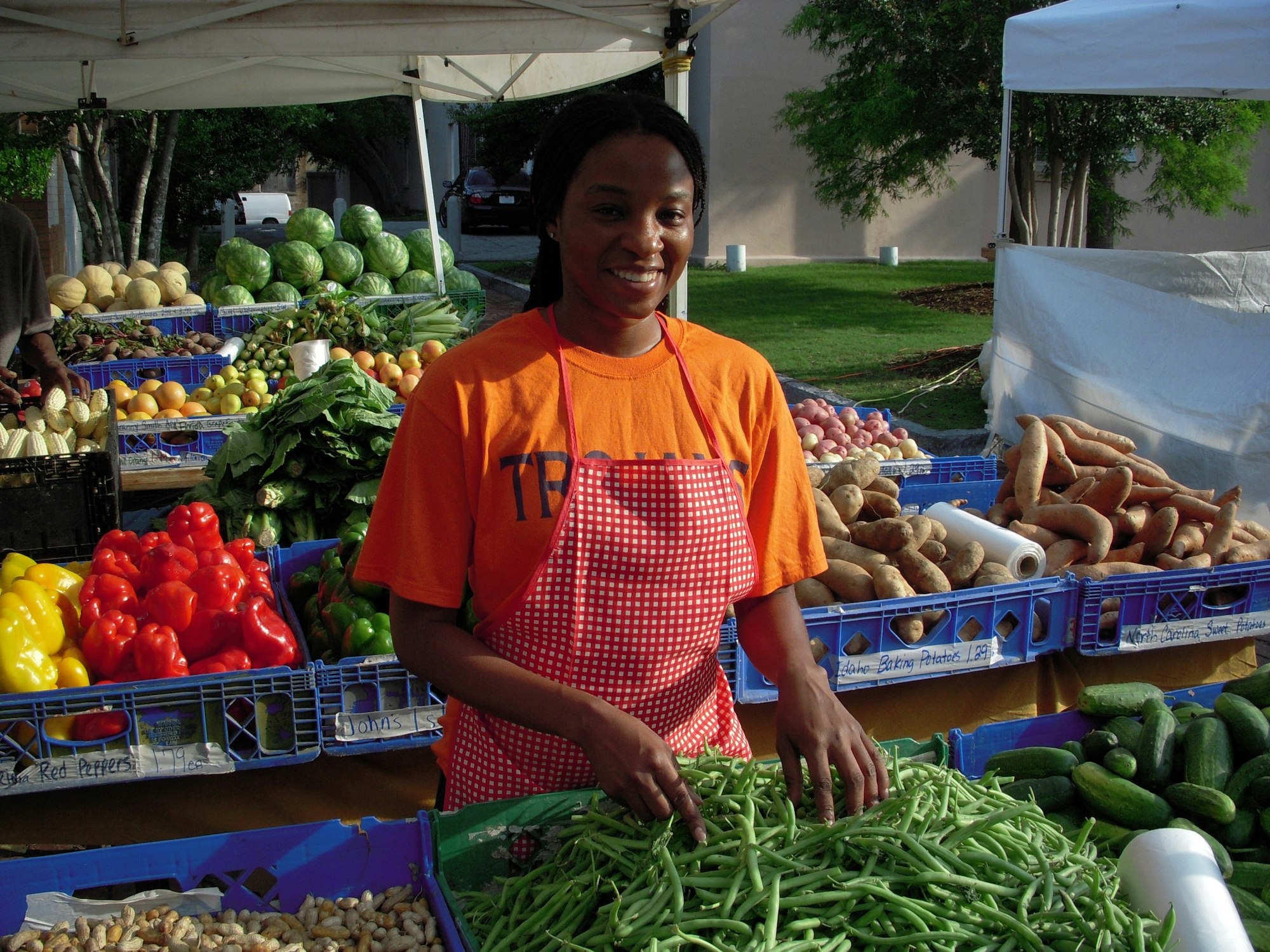 A woman in an orange shirt and apron smiles behind a table of fresh vegetables at a market.