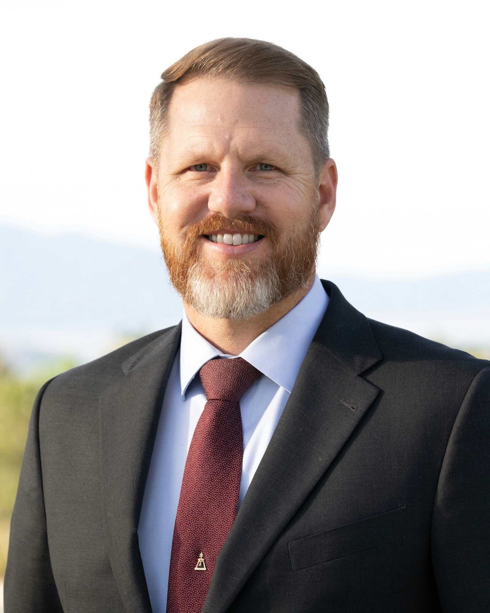 A man in a suit smiling, wearing a red tie with a pin.