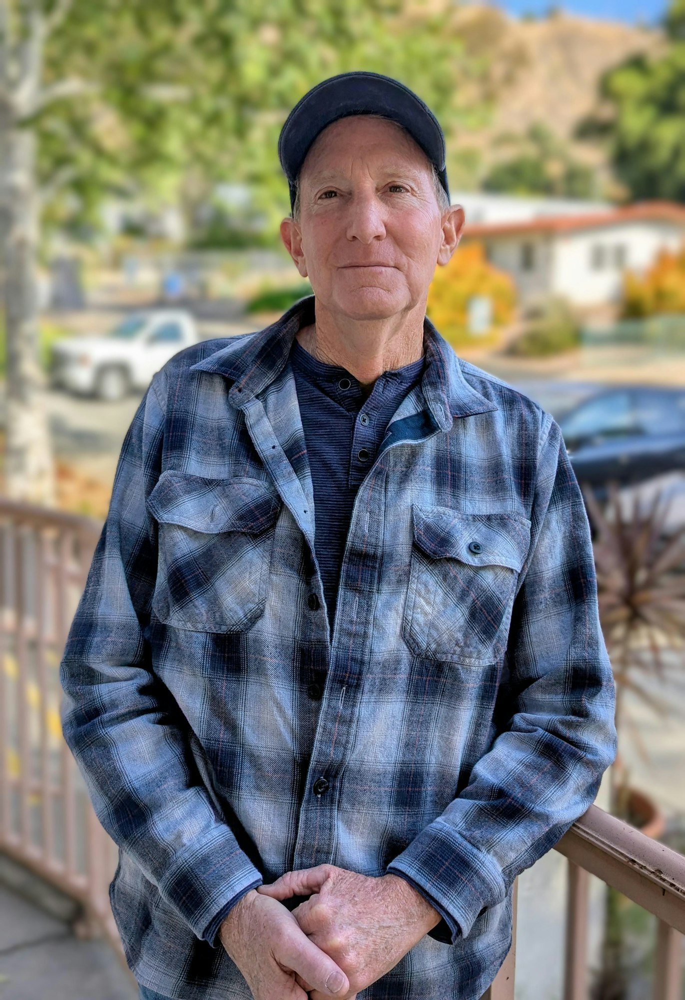 A man stands outside, wearing a plaid shirt and cap, with a blurred background of buildings and trees.