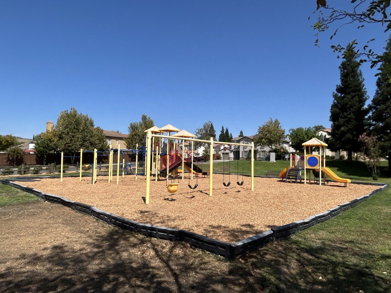 A colorful playground featuring swings, slides, and a playstructure, surrounded by grassy areas and trees under a clear blue sky.