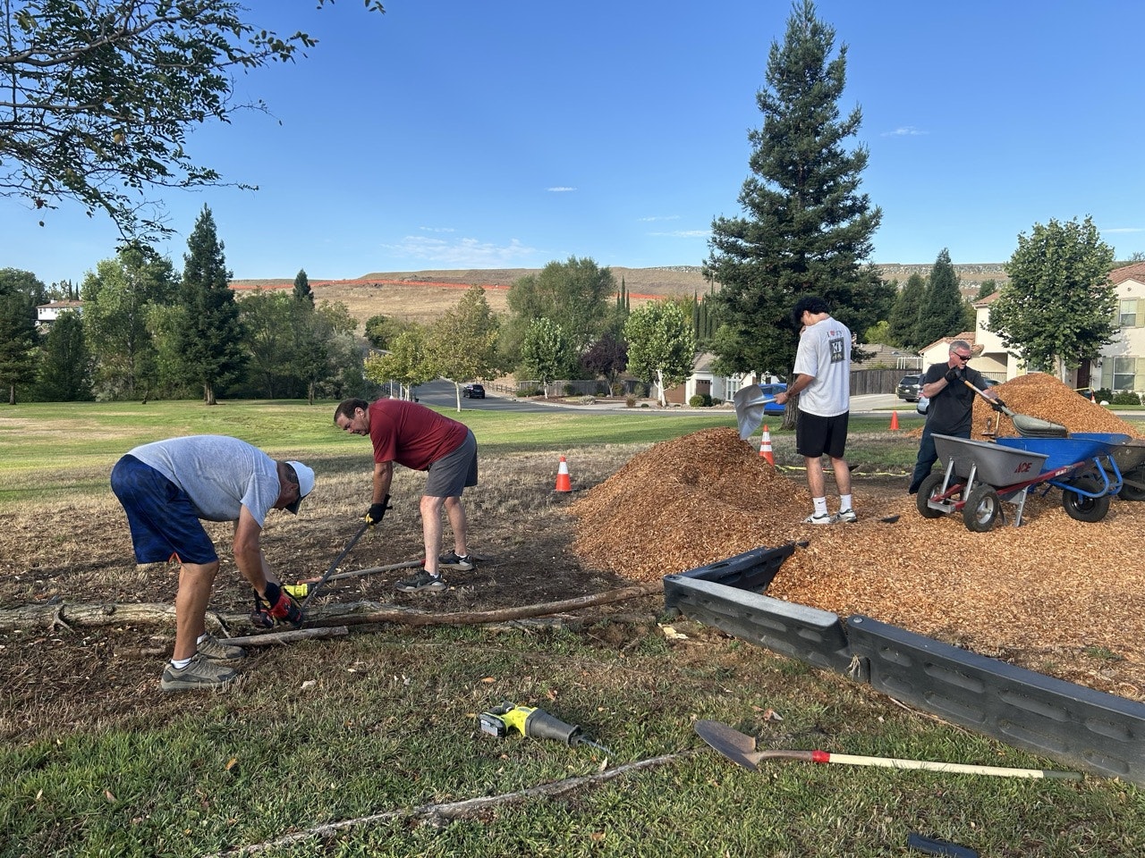 A group of people is working outdoors, using tools to clear and prepare an area with a pile of wood chips in a park setting.