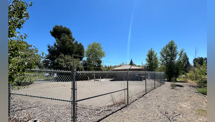 Chain link fence surrounds a concrete area with a small building. Trees and a clear blue sky in the background.