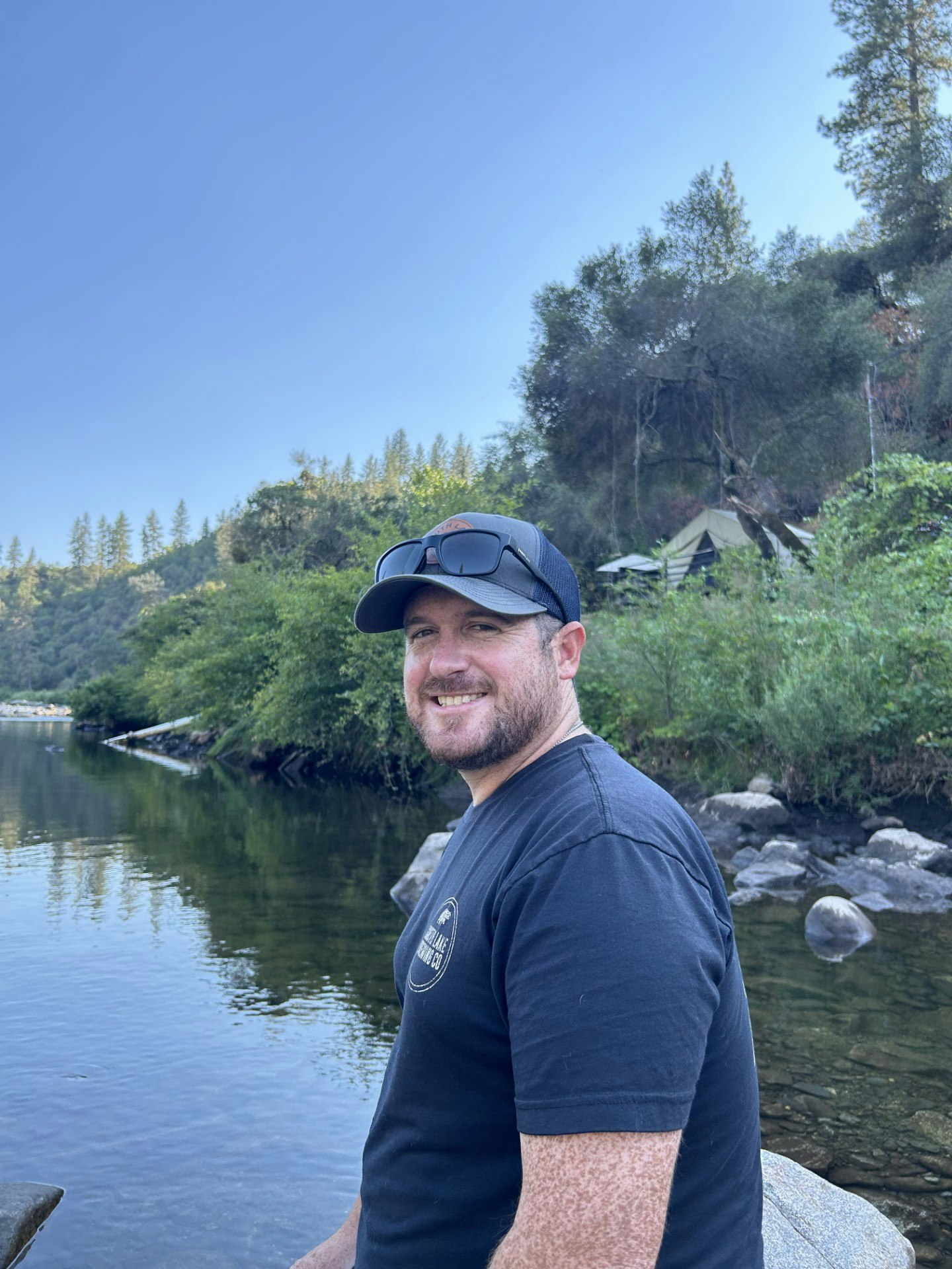 A man smiling near a river, surrounded by trees and rocks, with a tent in the background.