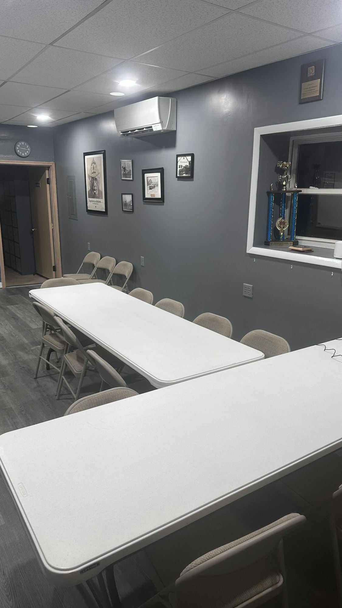 A meeting room with tables, chairs, a trophy cabinet, framed pictures, and a wall-mounted AC unit.