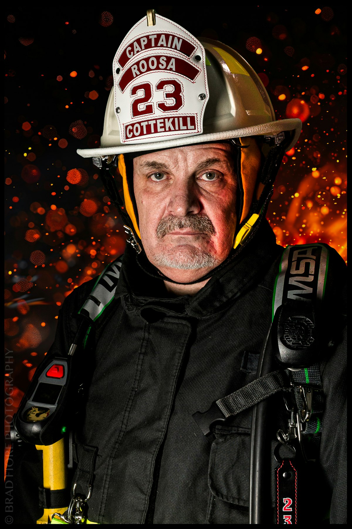 A firefighter in uniform, wearing a white helmet labeled "Captain Roosa 23 Cottekill," with orange sparks in the background.