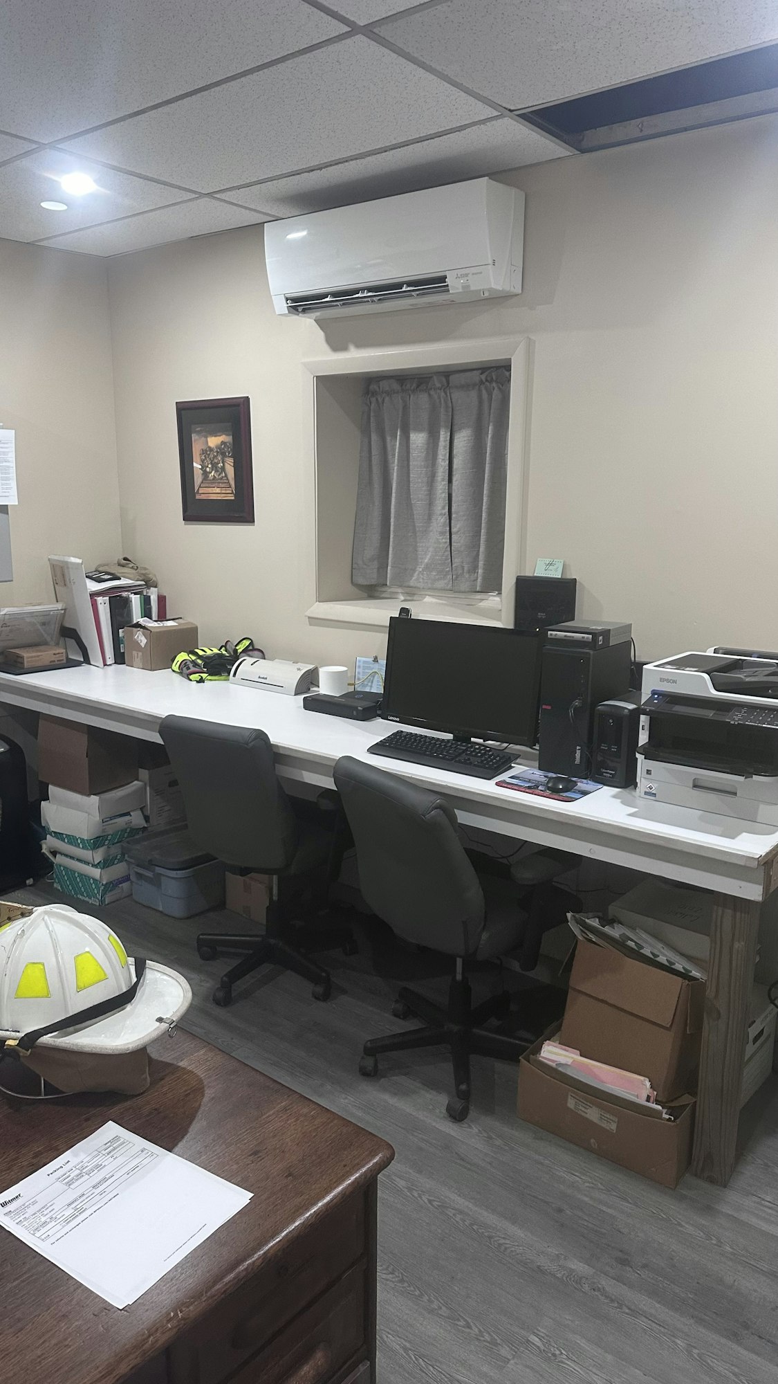 An office space with computers, printers, chairs, a desk with a hard hat, and an air conditioning unit.