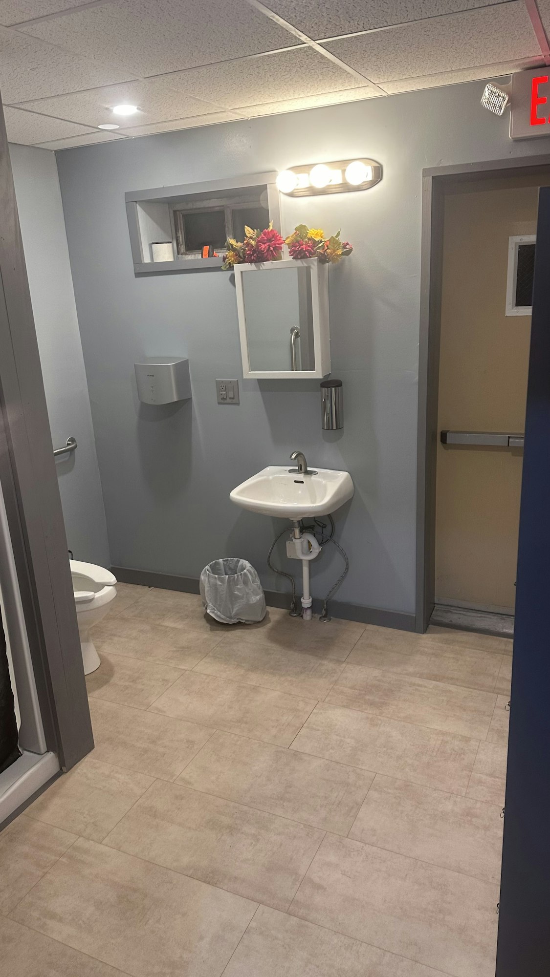 Public restroom interior with a toilet, sink, mirror, paper towels, trash can, and flowers on a shelf.