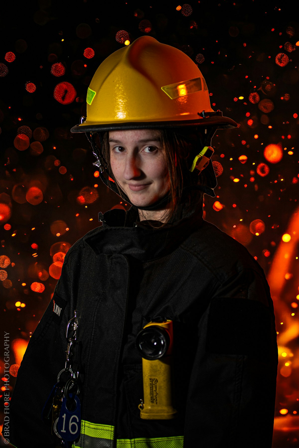 A person wearing a yellow firefighting helmet and black uniform with a flashlight attached, against a backdrop of glowing embers.