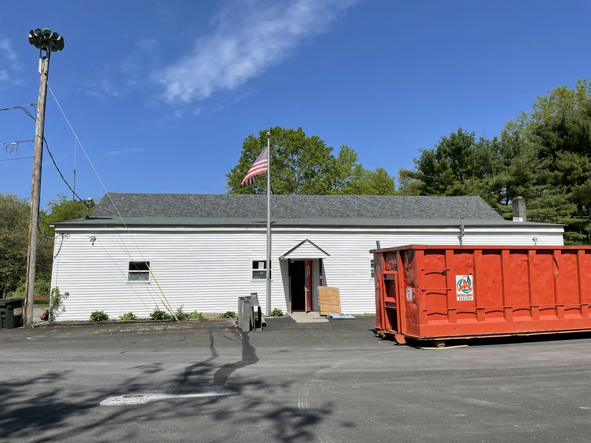 White building with an American flag, a large red dumpster, and a parking lot under a clear blue sky.