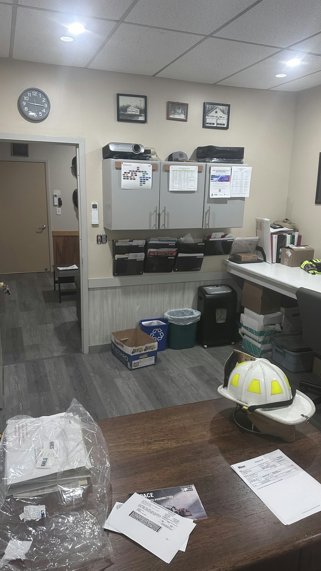 An office break room with a table, chairs, cabinetry, a clock on the wall, and a safety helmet on the table.
