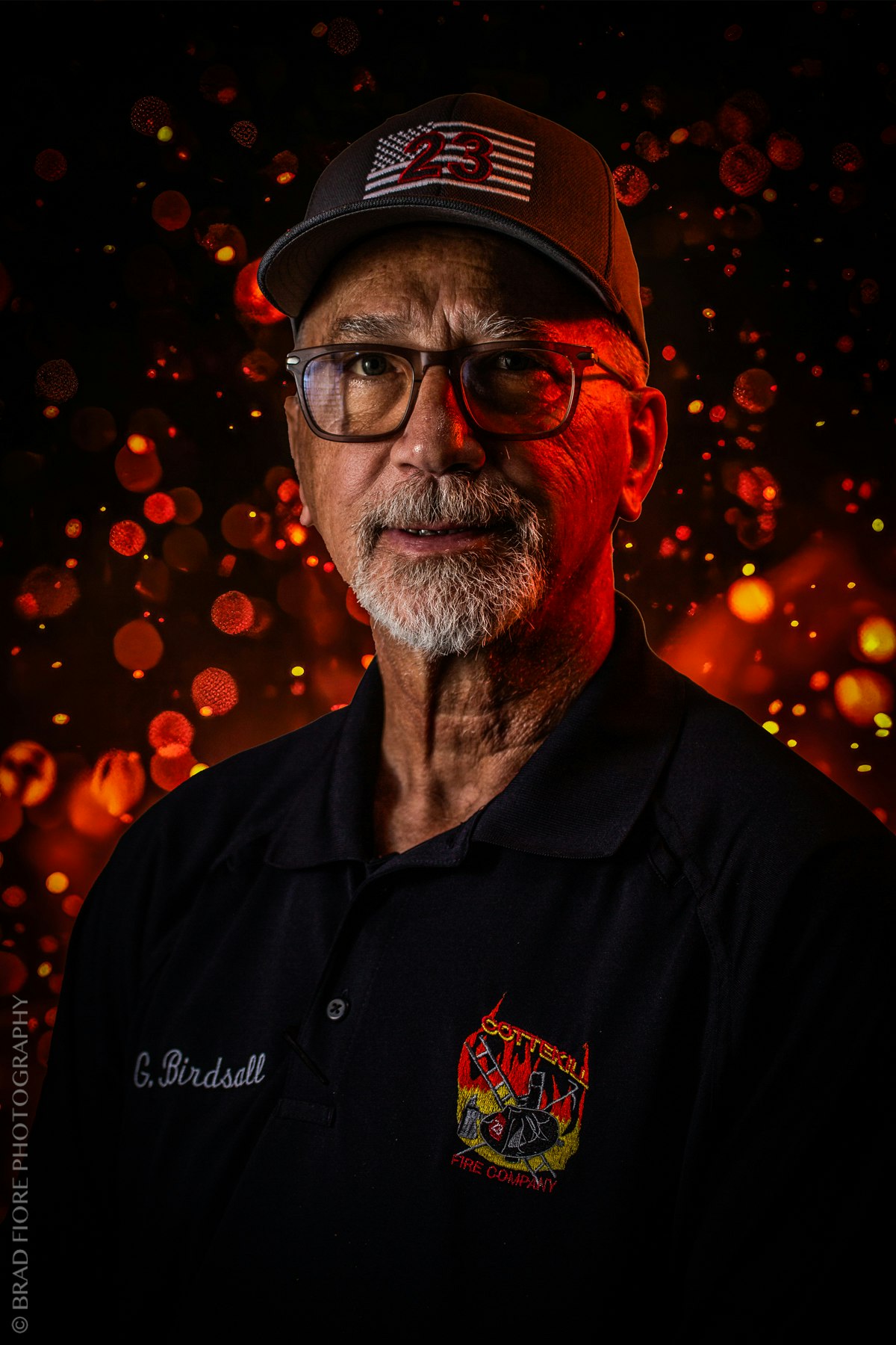 A man wearing glasses, a cap with "23" on it, and a black shirt with a "Cottekill Fire Company" logo, against an orange bokeh backdrop.