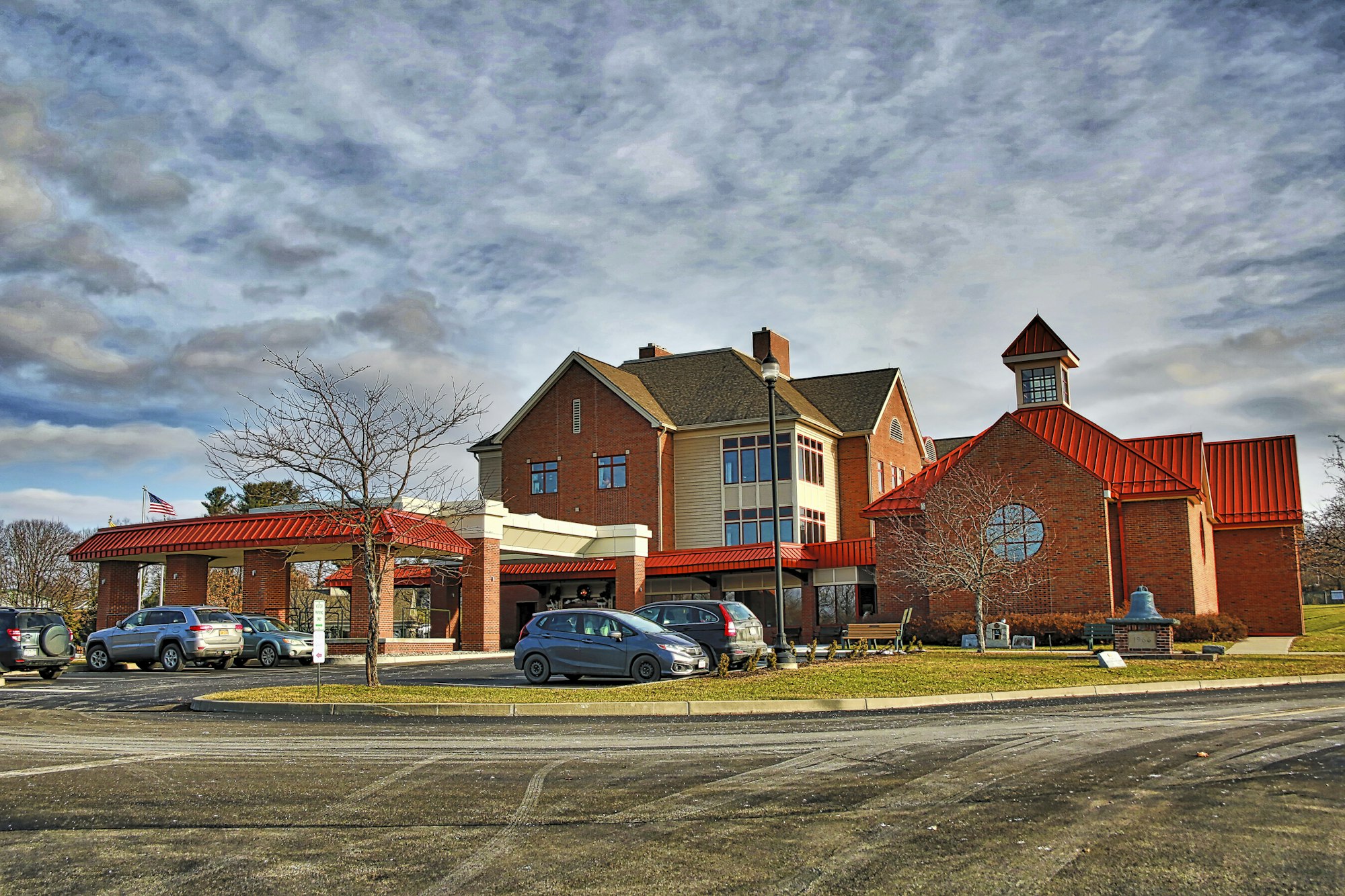 Brick building with a red roof, American flag, cloudy sky, and parked cars.