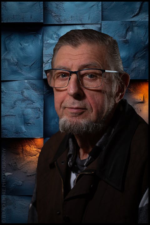 A man with glasses, a goatee, wearing a dark jacket, stands against a textured blue and orange geometric background.