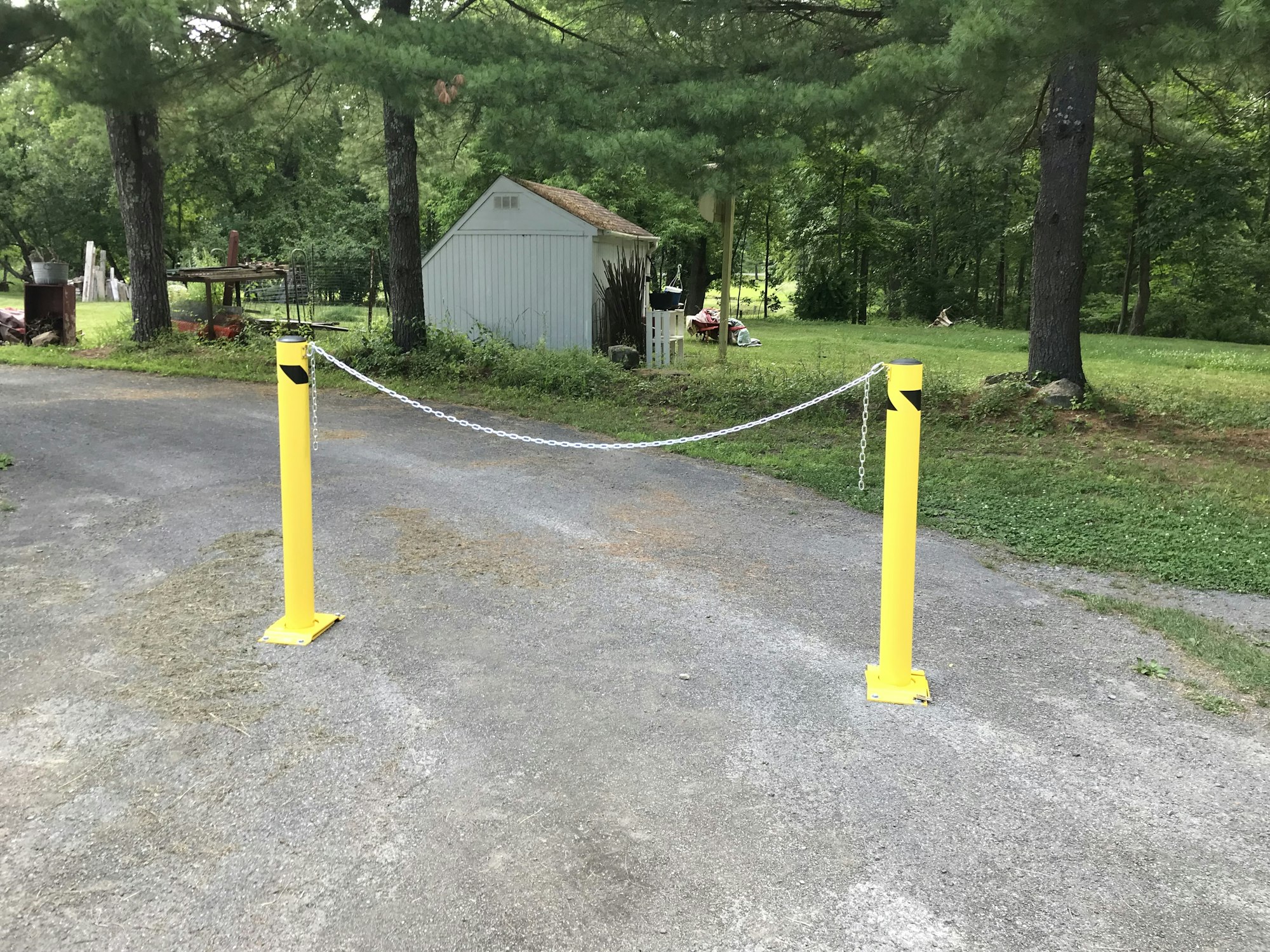 A driveway with yellow bollards connected by a chain, green trees, and a shed in the background.
