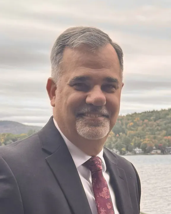 A man in a suit with a patterned tie smiles by a lake, surrounded by trees and hills in a scenic outdoor setting.