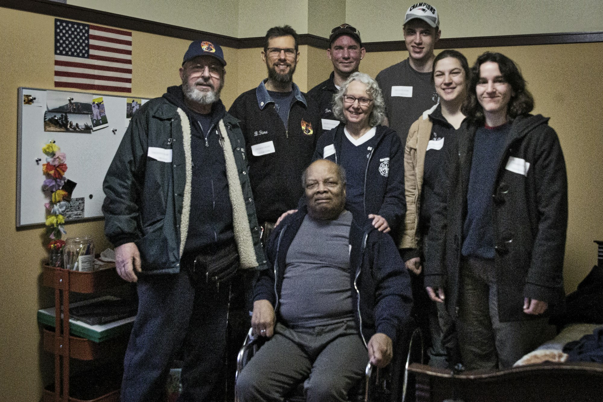 Group of people posing for a photo, one in a wheelchair, with an American flag in the background.