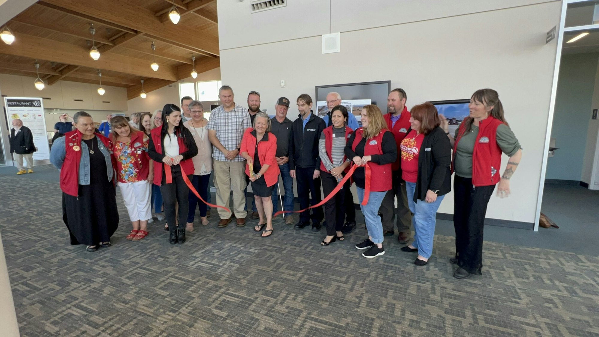 Group of people holding a red ribbon in a ceremonial event, likely a ribbon-cutting ceremony. They're indoors, smiling, and celebrating.