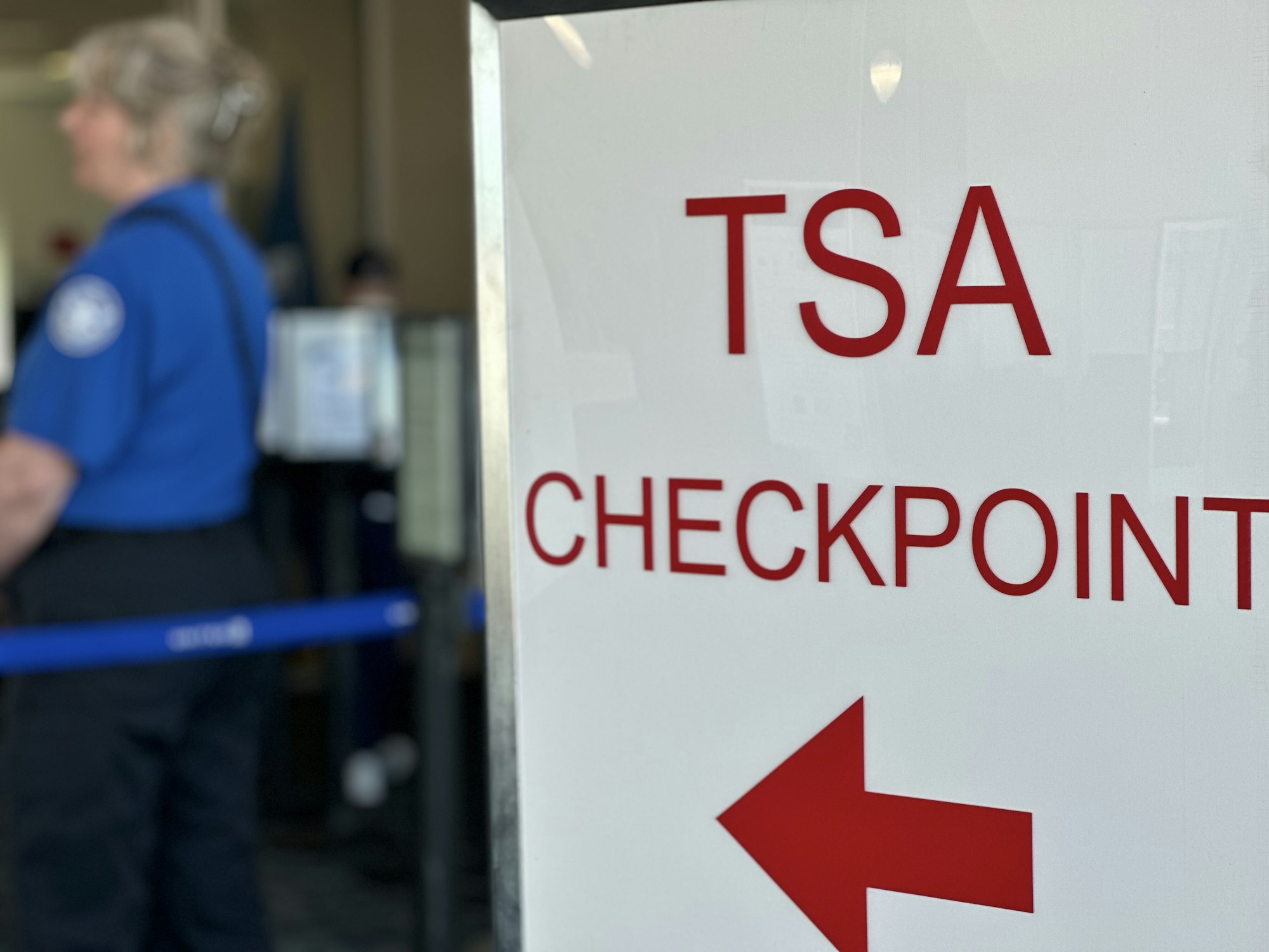 TSA checkpoint sign with an arrow, a security officer stands in the background.