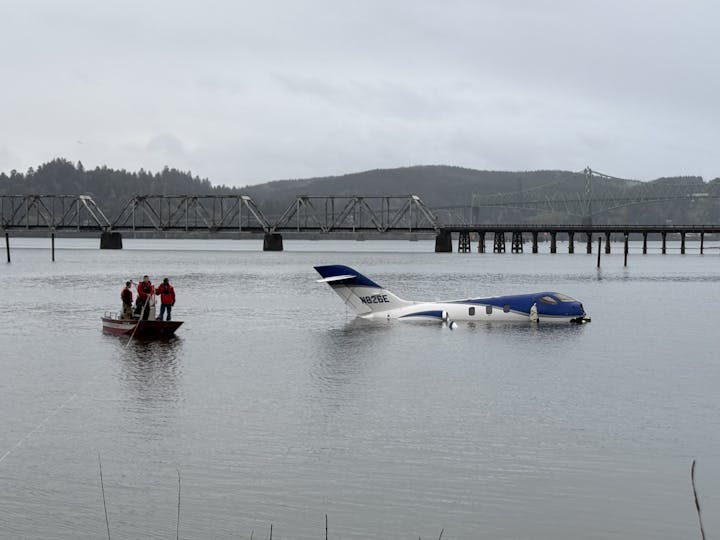 A small plane partially submerged in water with people on a boat nearby, near a bridge.