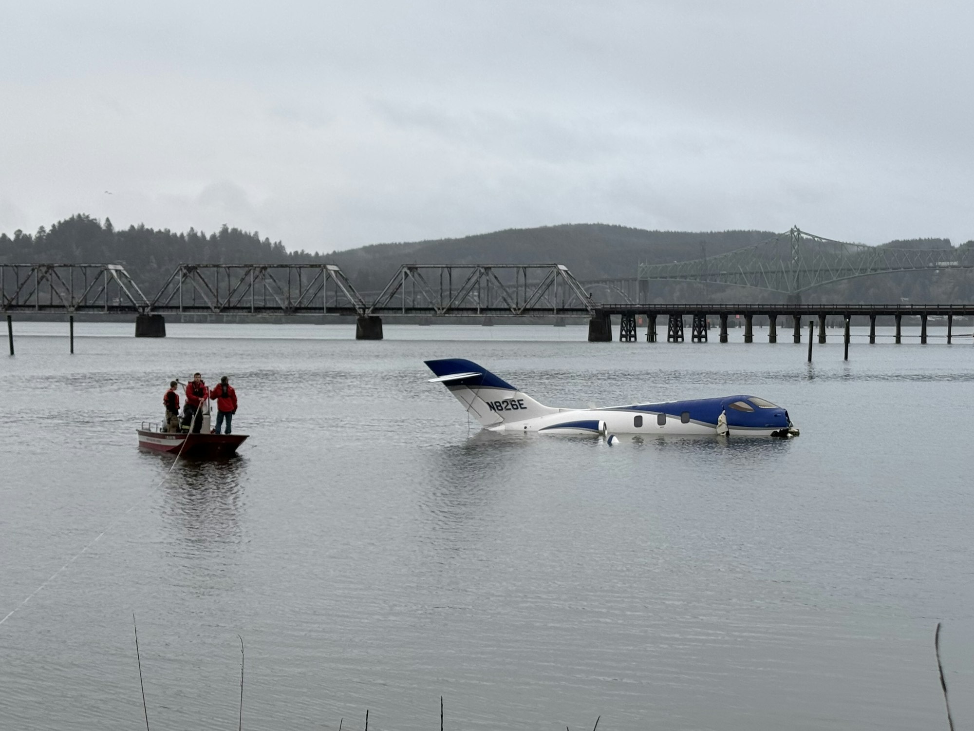 A small plane partially submerged in water with people on a boat nearby, near a bridge.