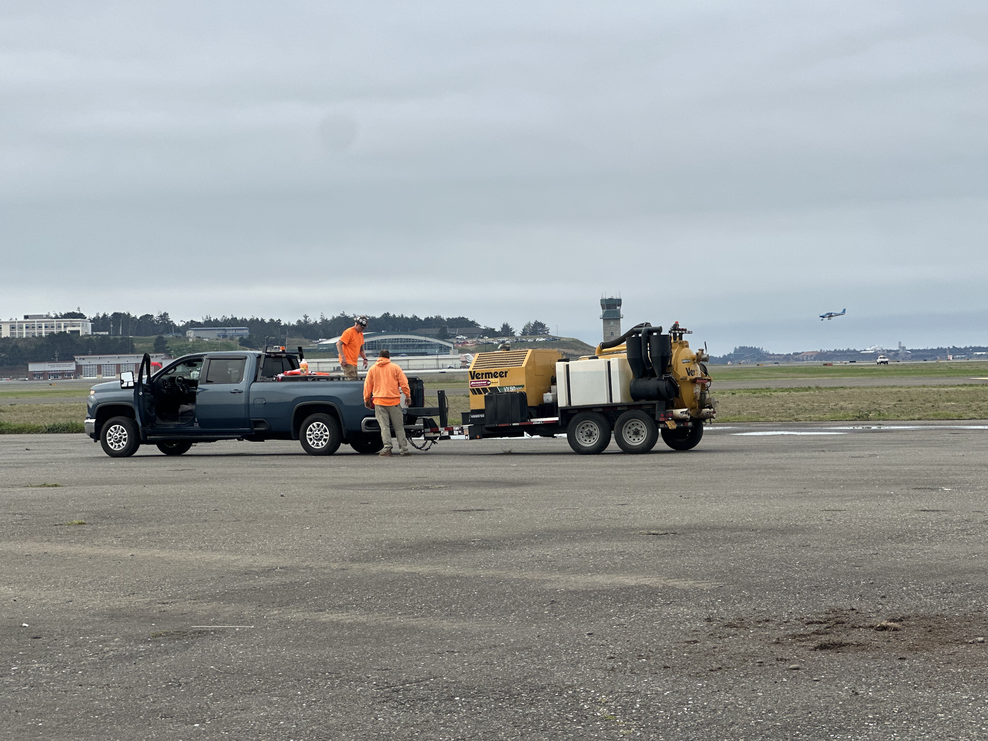 Truck and equipment with workers in fluorescent jackets on an airfield. An airplane is seen taking off in the background.