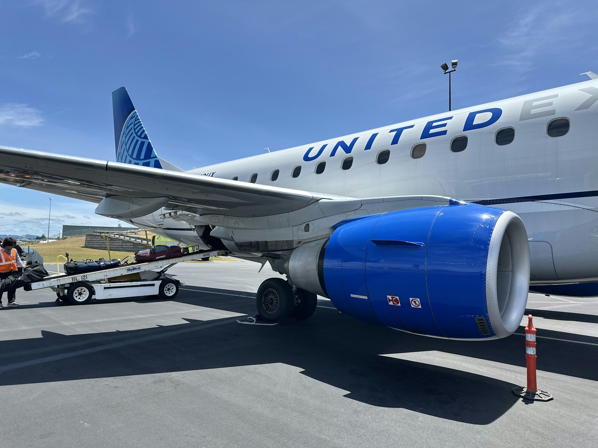 An aircraft with blue engine and tail is parked on the tarmac, with luggage being loaded via a ramp.