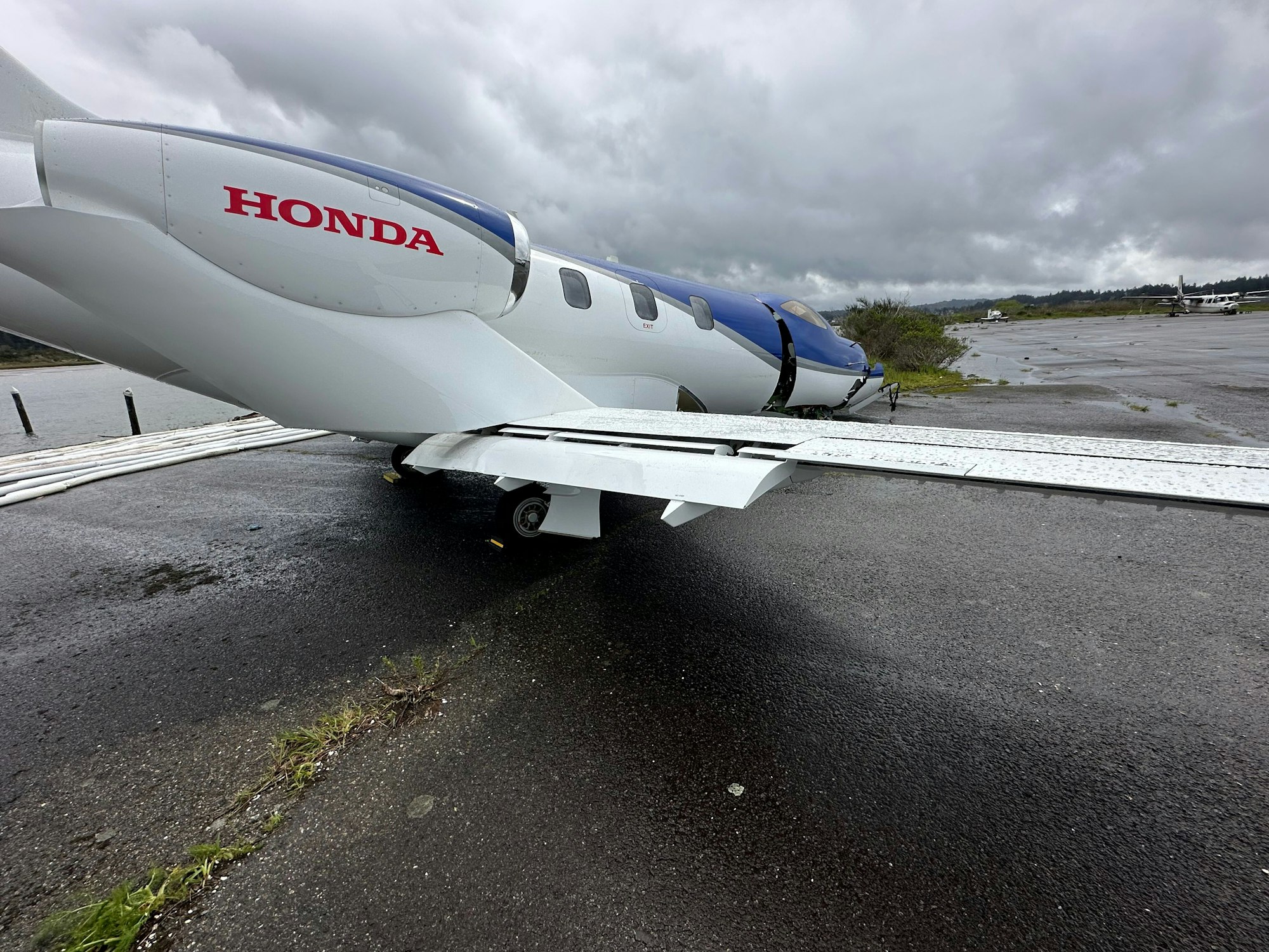 A small white jet with "HONDA" on the engine is parked on a wet runway under a cloudy sky.
