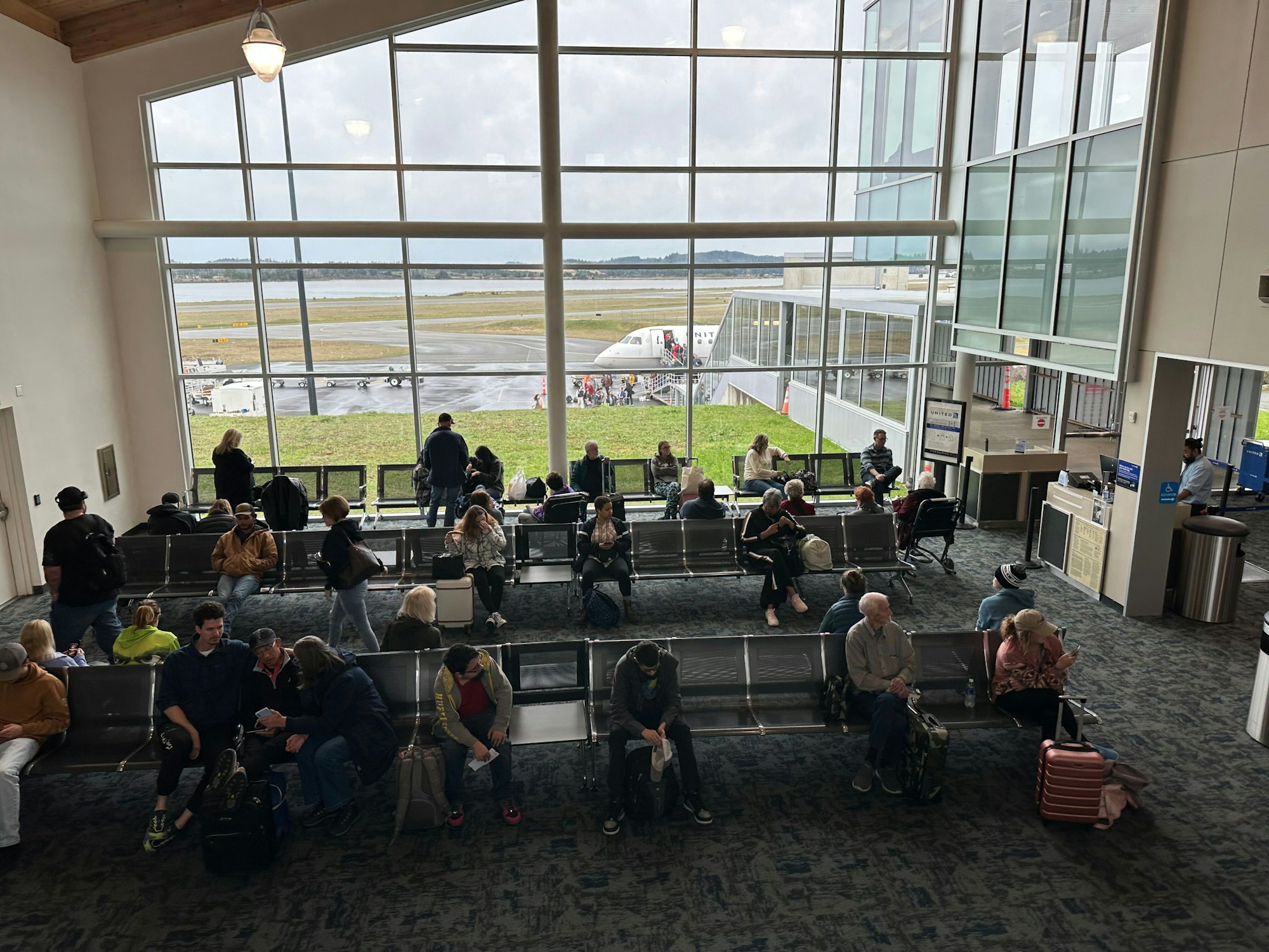 People seated in an airport lounge, waiting near large windows with a view of the tarmac and a plane outside.
