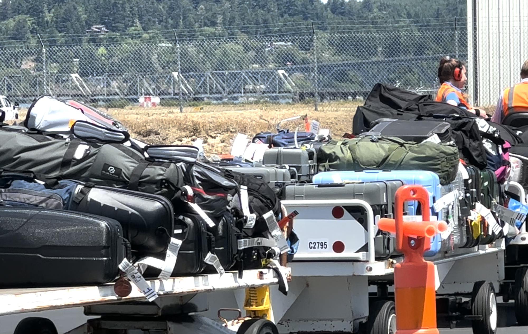 The image shows a stack of luggage on carts at an airport, with workers in safety vests managing the bags.
