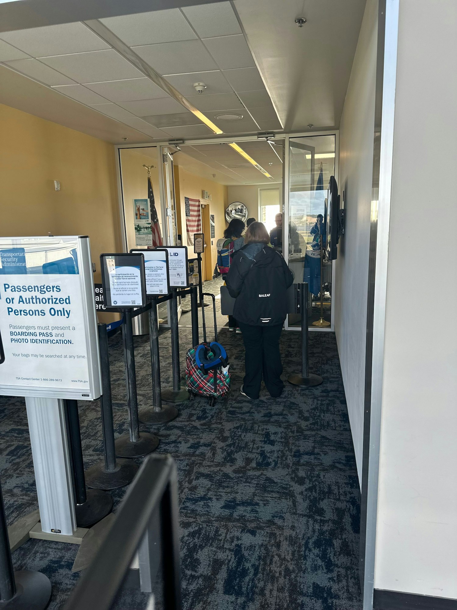 An airport security checkpoint with signs for authorized persons, passengers waiting, and a visible American flag.